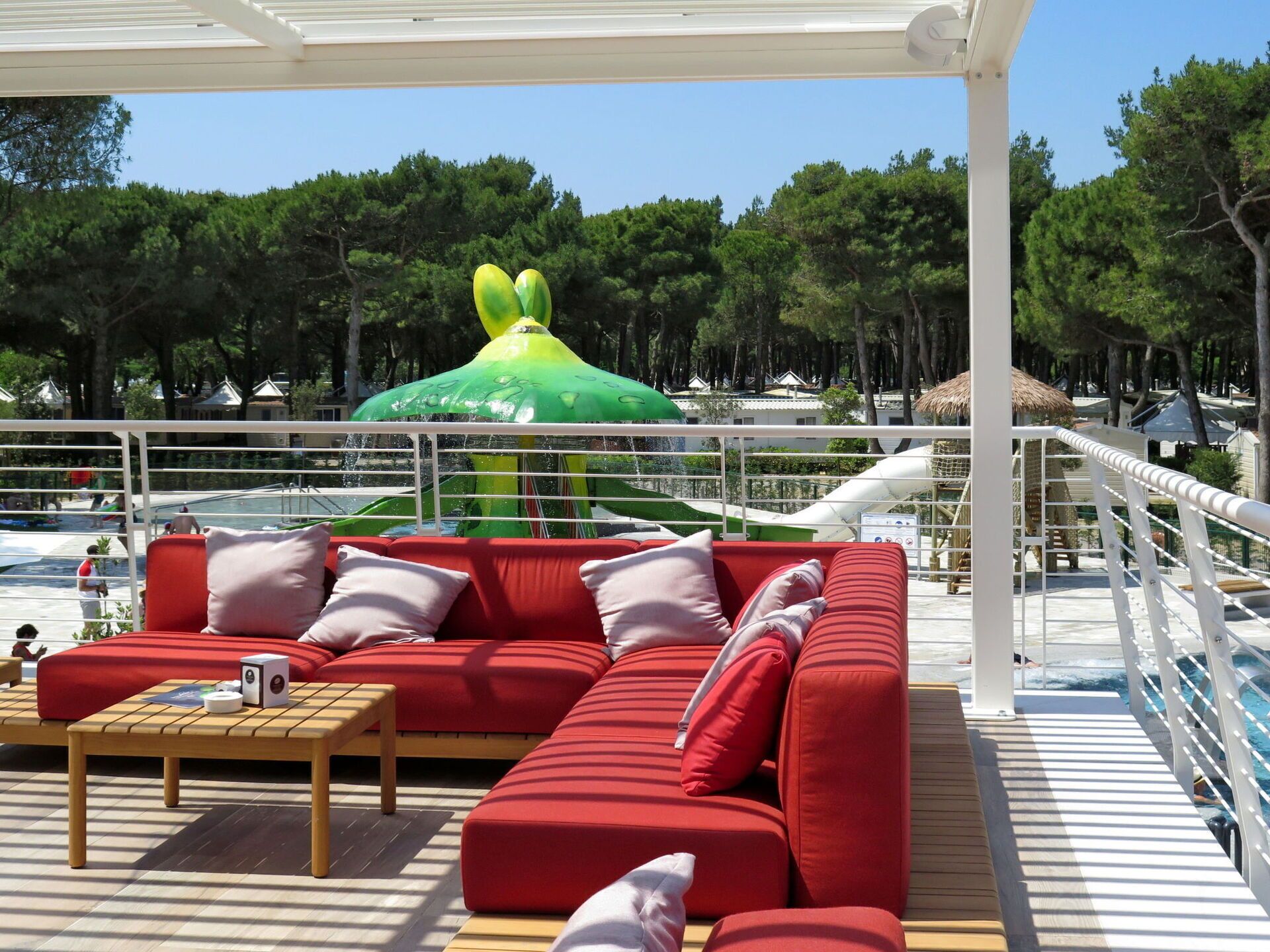 A covered outdoor seating area with a red sofa and a wooden table, overlooking a pool and a water slide.