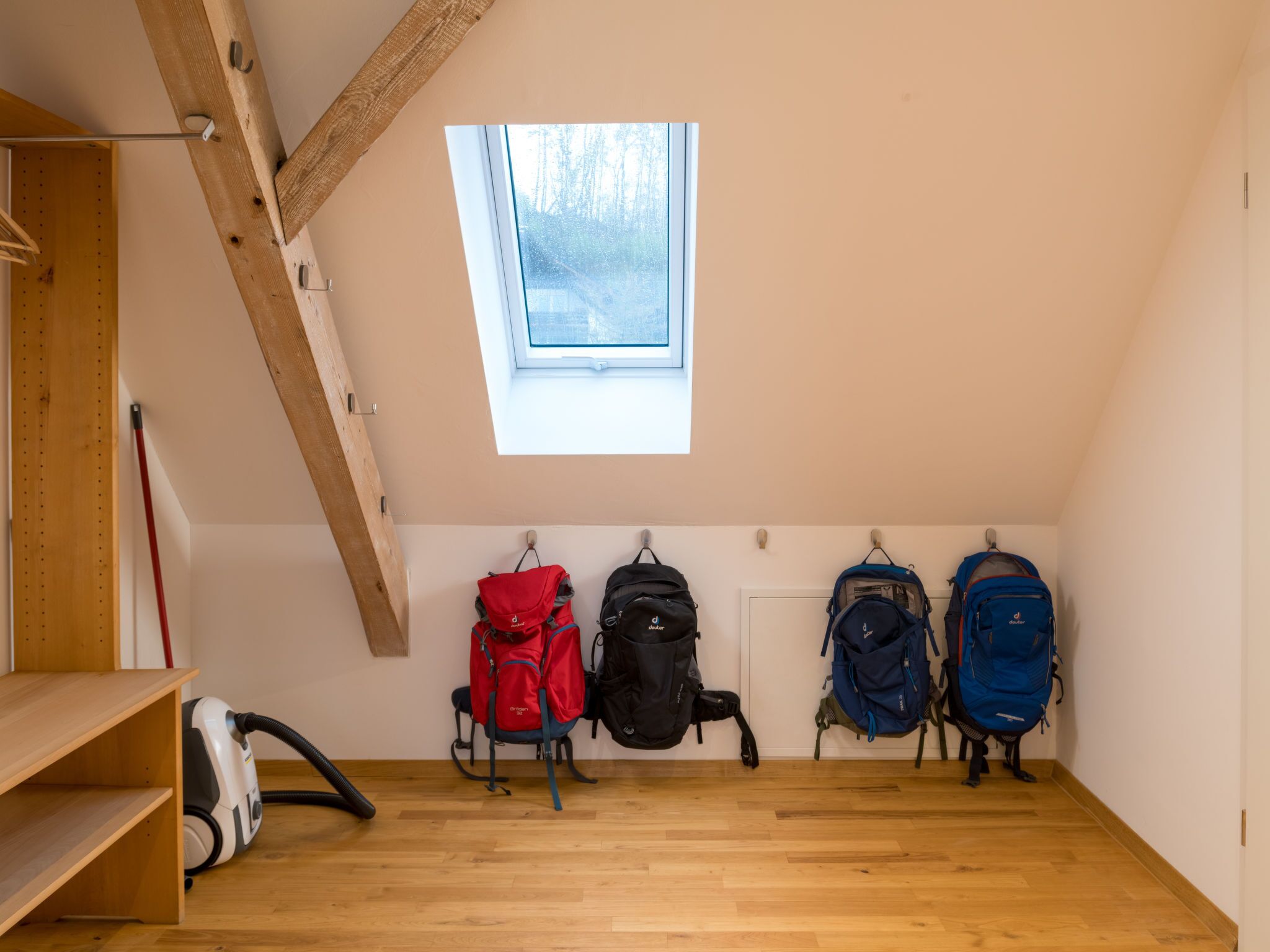 A room with a sloped ceiling, wooden beams, a skylight, and four backpacks hanging on the wall.