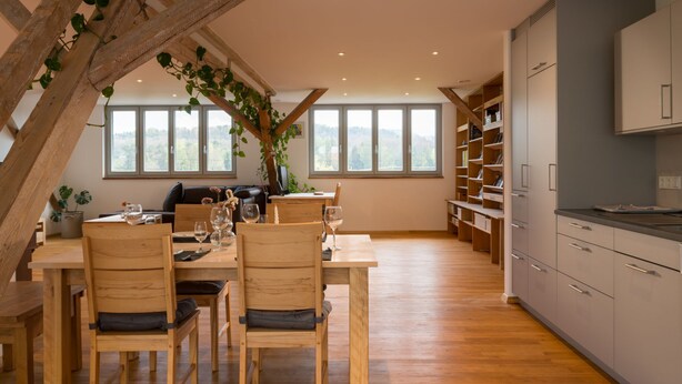 Una cocina moderna con vigas de madera, un comedor con mesa y sillas, y vistas al exterior a través de grandes ventanales.
