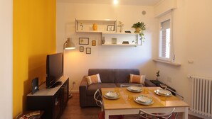 A compact living space with a sofa, a dining table set for two, a TV, and shelves with decorative items. - The Yellow Petal (Verona)