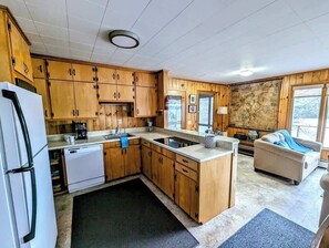 A compact kitchen with wooden cabinets, a dishwasher, a refrigerator, a sink, and a small seating area with a sofa and a window.