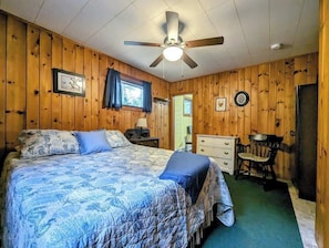 Una habitación con una cama, ventilador de techo, cómoda, silla y puerta.