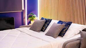 Een opgemaakt bed met witte lakens en meerdere kussens, een klein nachtkastje met een lamp, en een bloempot met een plant.