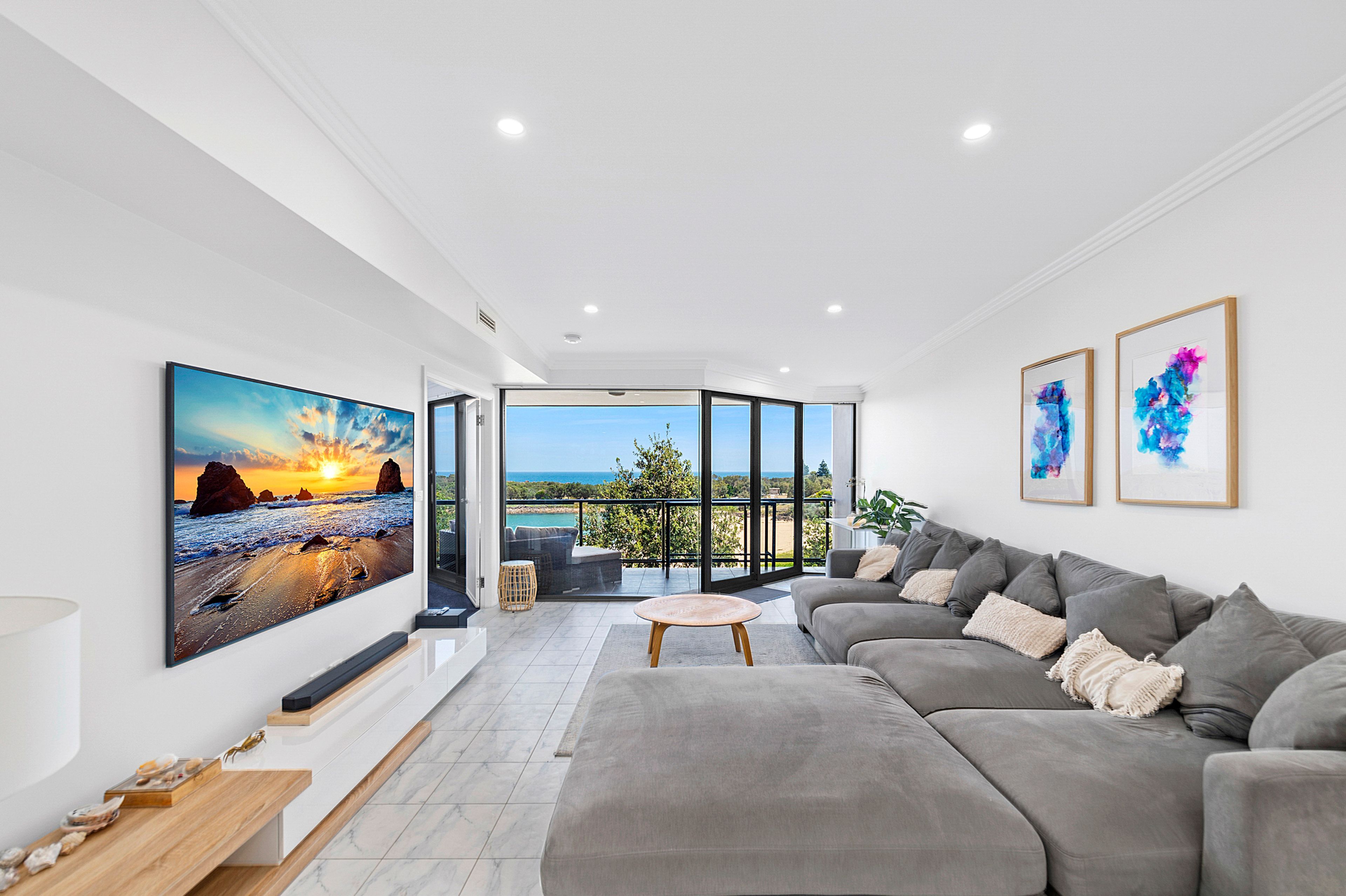 Un salón moderno con un televisor de pantalla plana grande, un sofá gris y vistas al mar a través de puertas de vidrio.