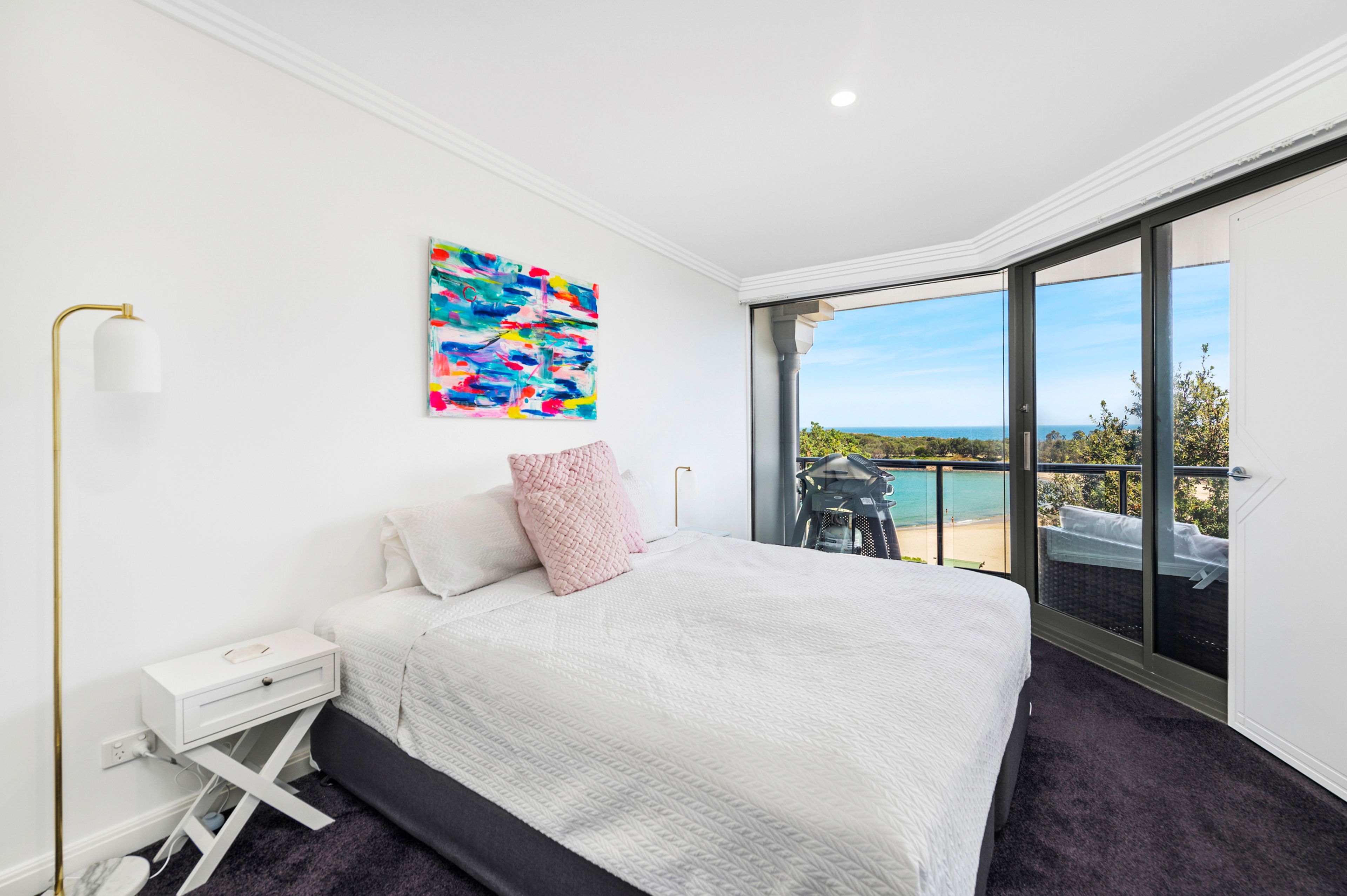 A bedroom with a bed, a nightstand, a lamp, a colorful abstract painting, and a balcony with a view of the sea.