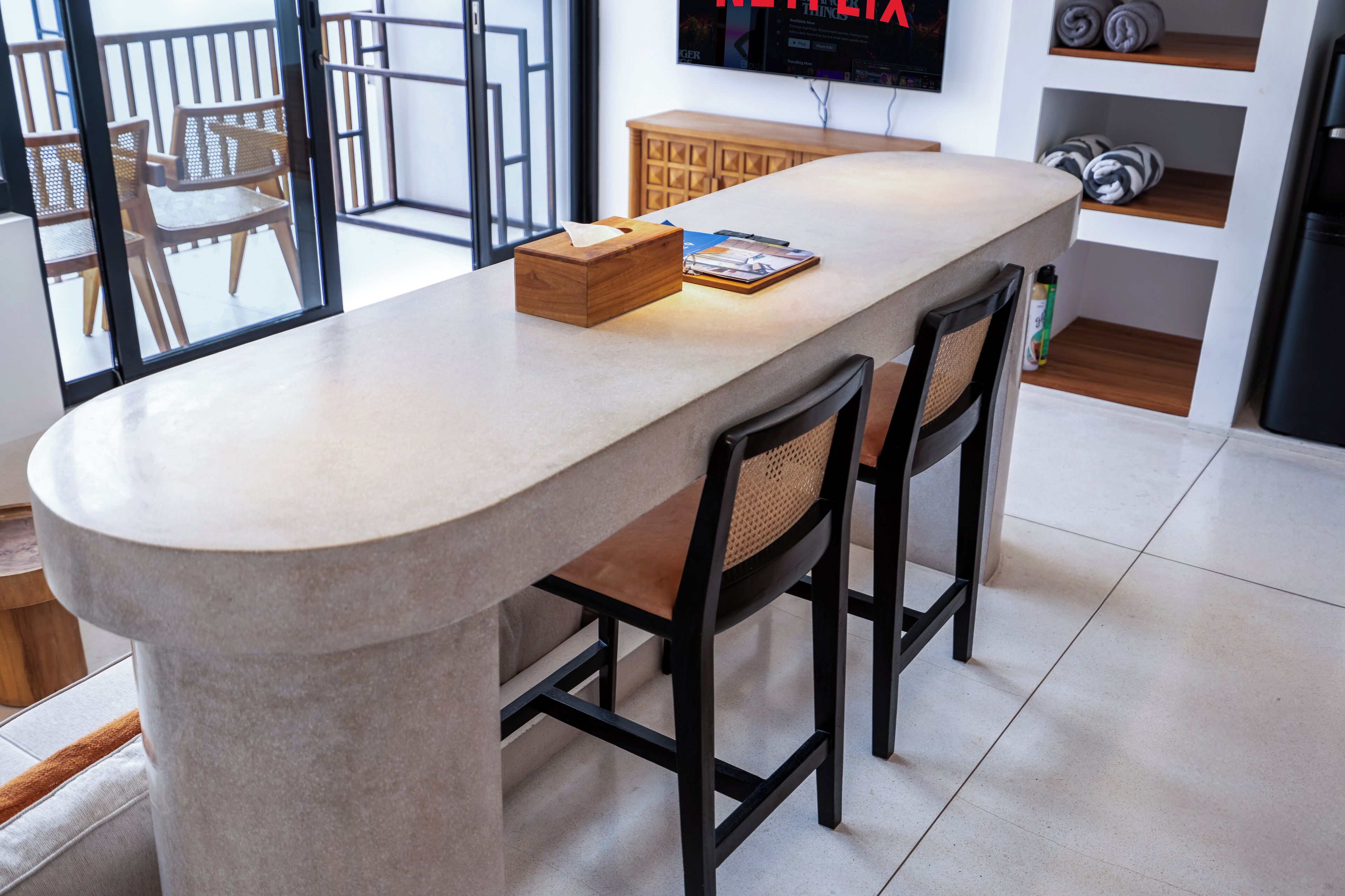 A modern kitchen with a curved island, two bar stools, a wooden cabinet, and a television mounted on the wall.