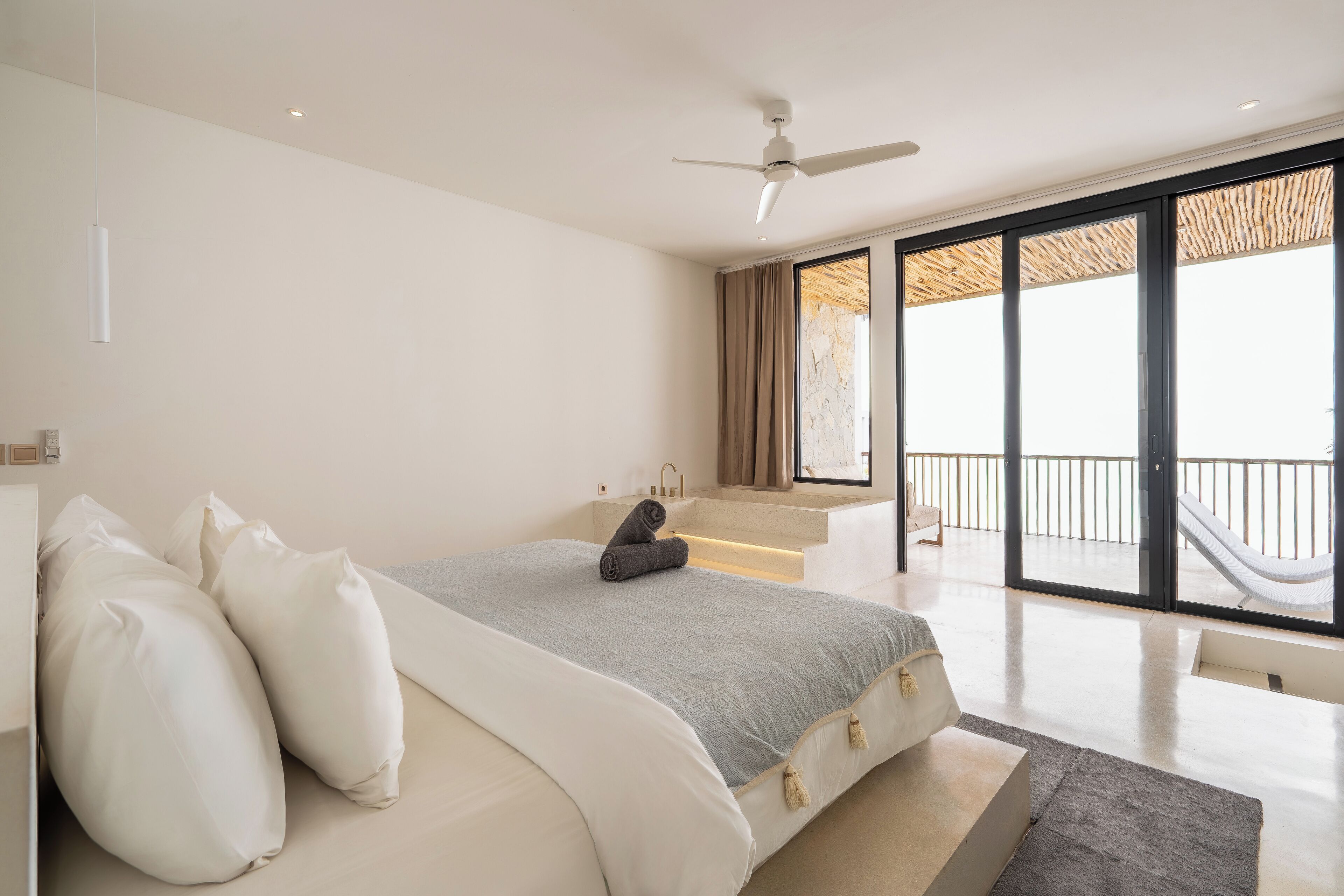 Un dormitorio con cama, ventilador de techo, ventana grande y vista a un balcón.
