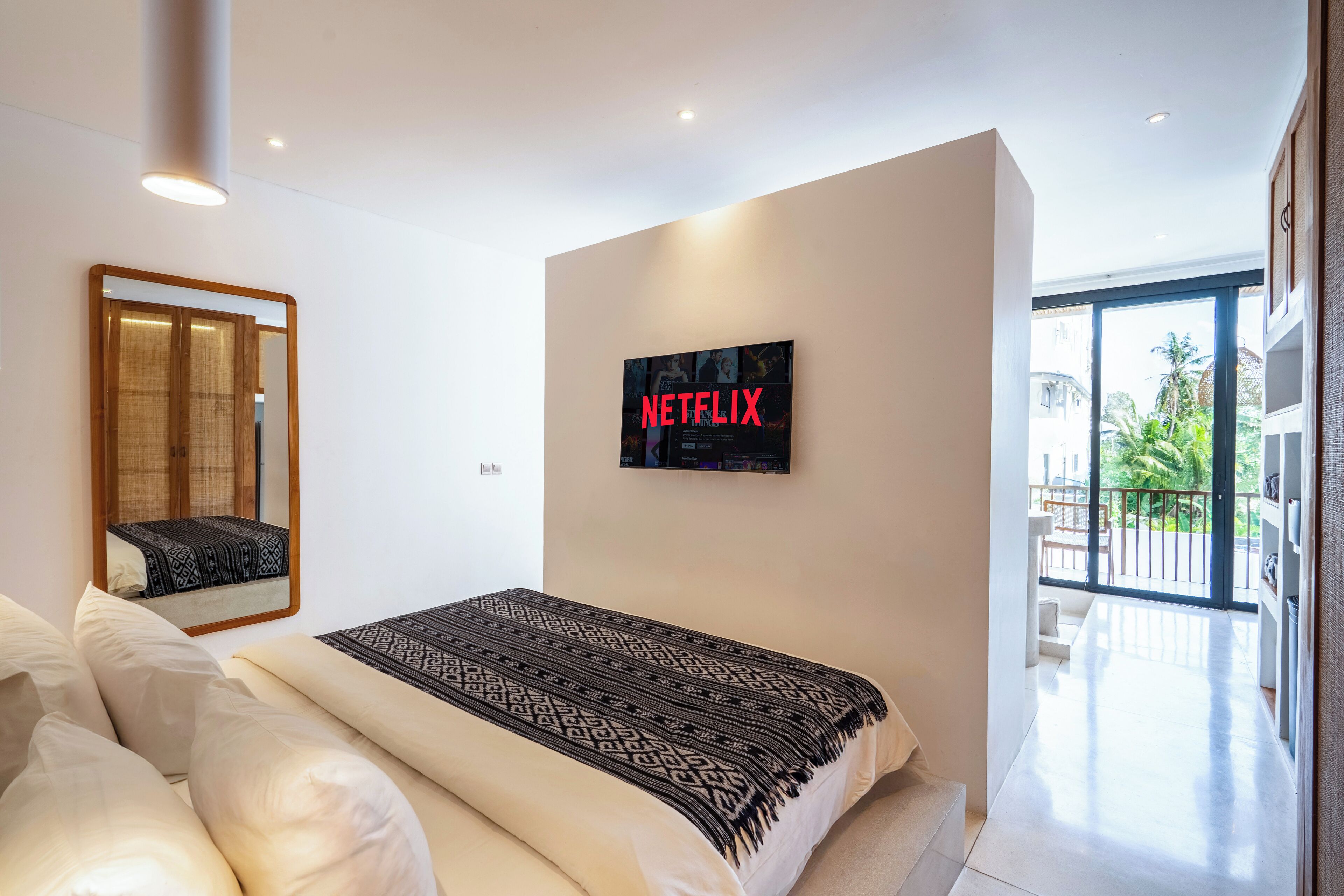 A modern bedroom with a large bed, a wall-mounted TV displaying 'NETFLIX', and a balcony with a view of palm trees.