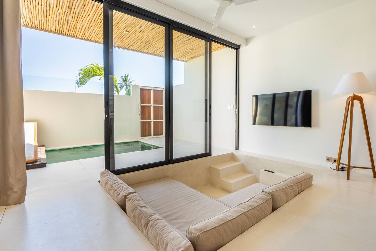 Un salon moderne doté d’une grande porte-fenêtre coulissante donnant sur une piscine, d’un téléviseur à écran plat fixé au mur et d’un lampadaire.