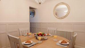 Une salle à manger avec une table ronde, des chaises blanches, un lustre, un miroir et un saladier rempli de fruits.