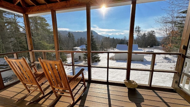 A wooden deck with rocking chairs and a view of a snowy landscape through large windows. - 11G Quintessential New England Cottage with Fantastic Views and Dog Friendly! (Sugar Hill)
