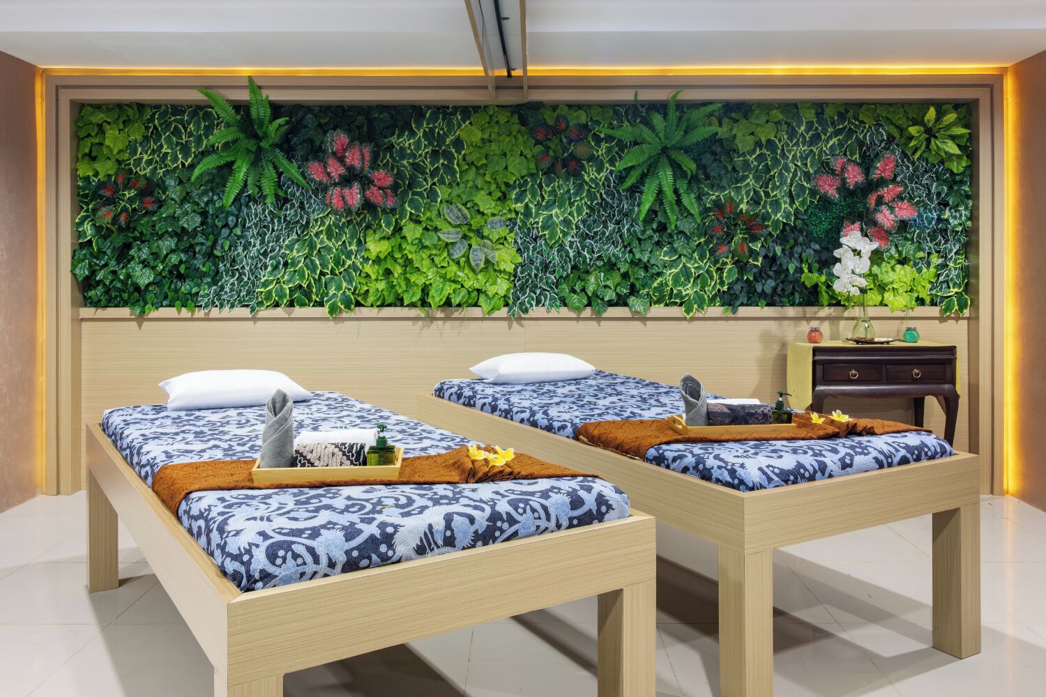 A hotel room with two beds, a wooden headboard, a wall-mounted plant display, and a small bedside table with a vase.