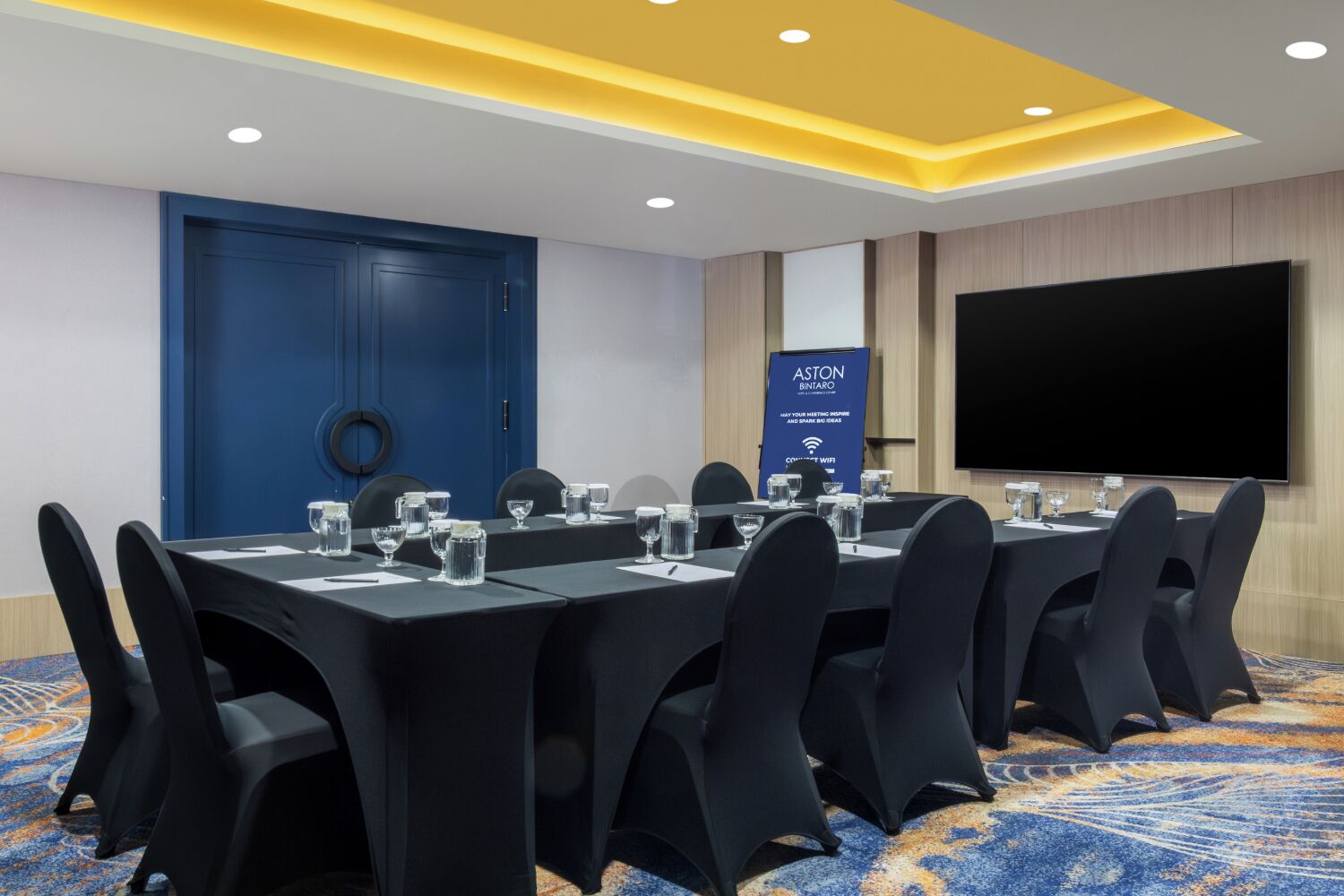 A conference room with a long table, chairs, a large screen, and a blue door.