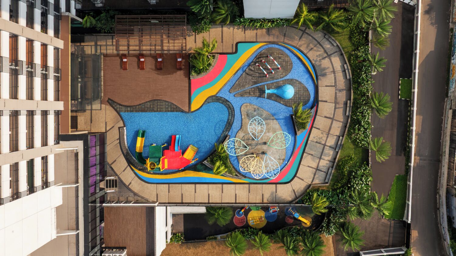 Vista aèria d'un parc infantil amb diverses estructures de joc, envoltat de palmeres i un edifici.