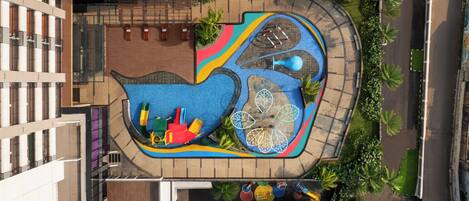 Vista aérea de un parque infantil con diversas estructuras de juego, rodeado de palmeras y un edificio.