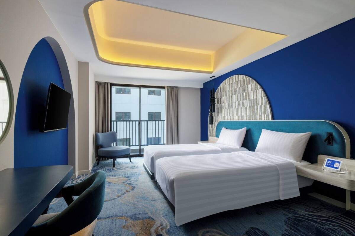 A hotel room with two beds, a desk, a chair, a TV, and a balcony with a city view.