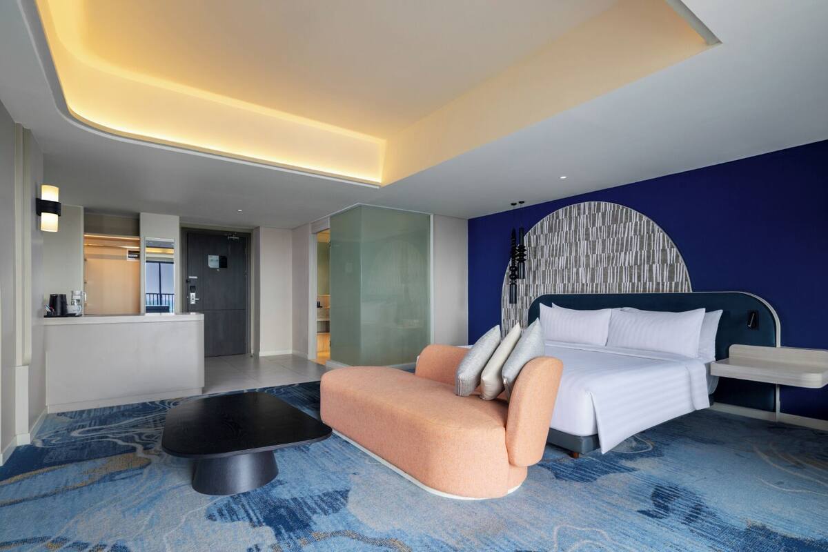 A modern hotel room with a bed, a sofa, a coffee machine, and a reception area.