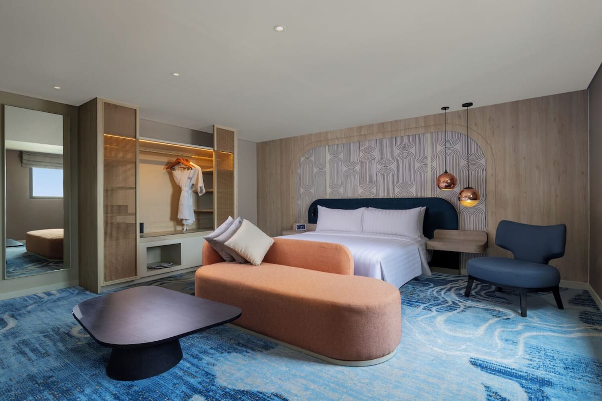 A modern hotel room with a large bed, a seating area with a sofa and armchair, a walk-in closet, and a patterned carpet.