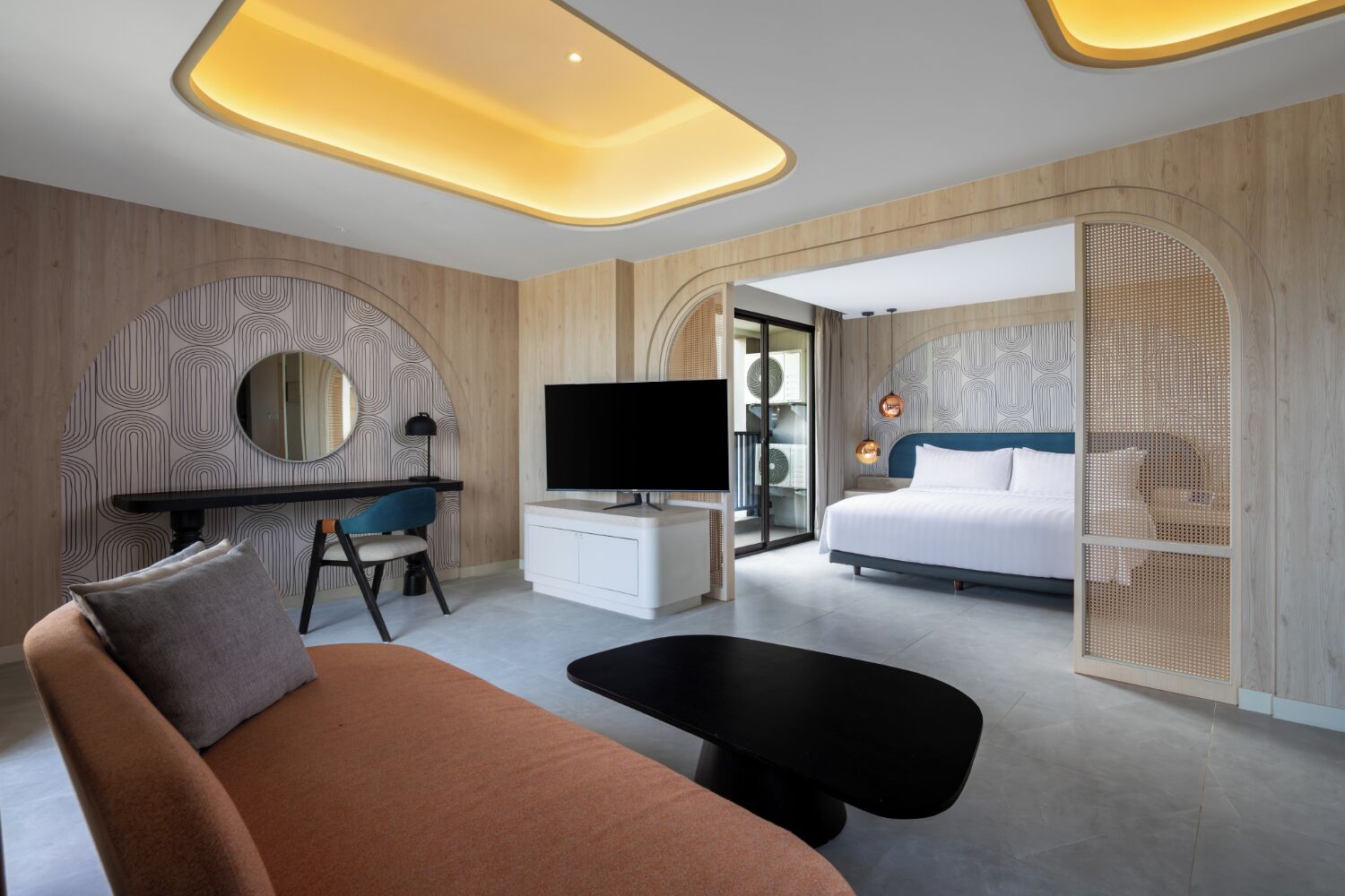 A modern hotel room with a sofa, a desk with a TV, a bed, and a balcony with a view.