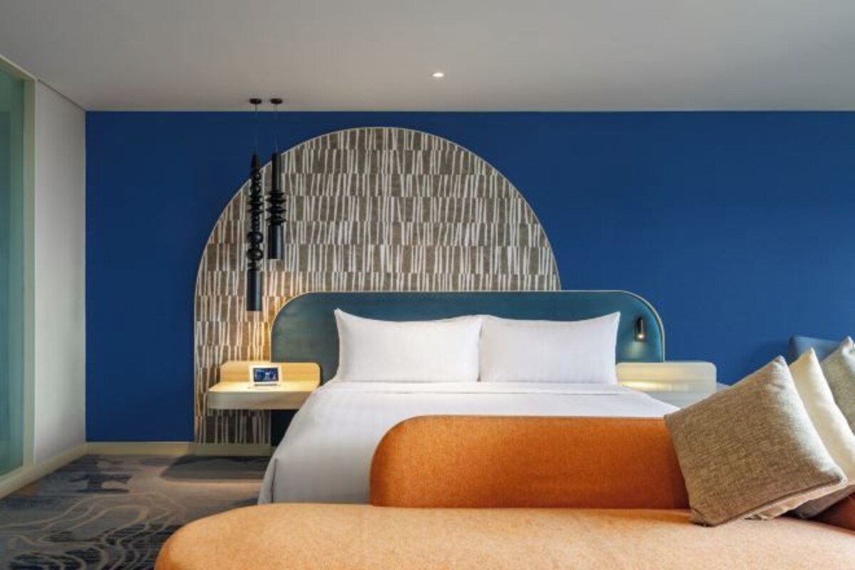 A modern hotel room with a large bed, a curved wall design, and a nightstand with a lamp.