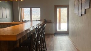 Une cuisine moderne avec un bar en bois et des tabourets de bar, de grandes fenêtres et une vue sur l’extérieur.