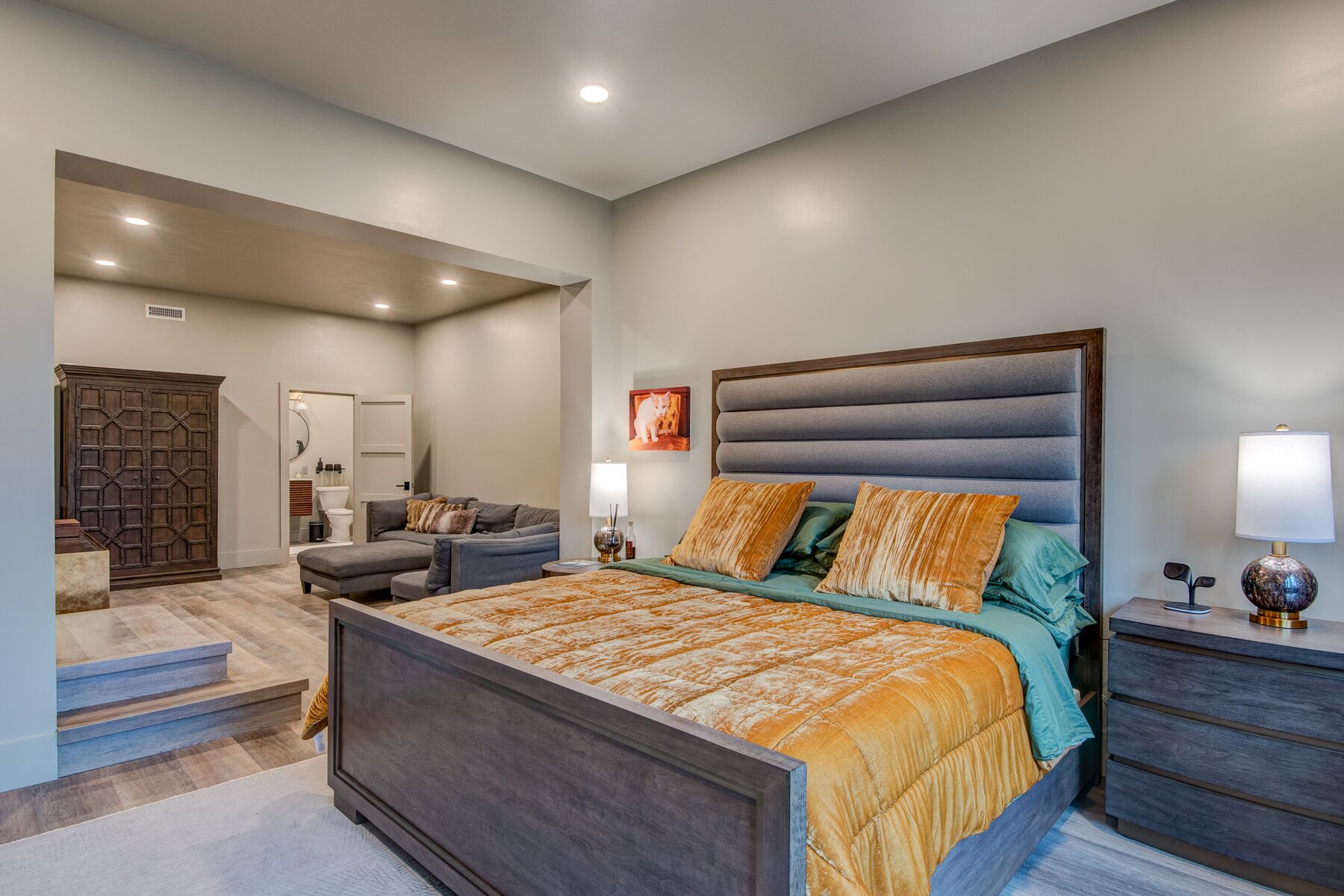 A bedroom with a large bed, a nightstand, a dresser, a wardrobe, and a bathroom visible in the background.