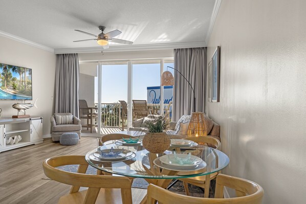A spacious living room with a glass-top dining table, comfortable seating, a ceiling fan, and a view of the outdoors through large windows. - Destin West Gulfside 409 ~ Ocean Views ~ Free Beach Service! (Fort Walton Beach)