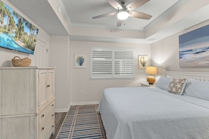 Un dormitorio con cama, ventilador de techo, televisor, ventana con persianas, lámpara y una concha decorativa en la pared.