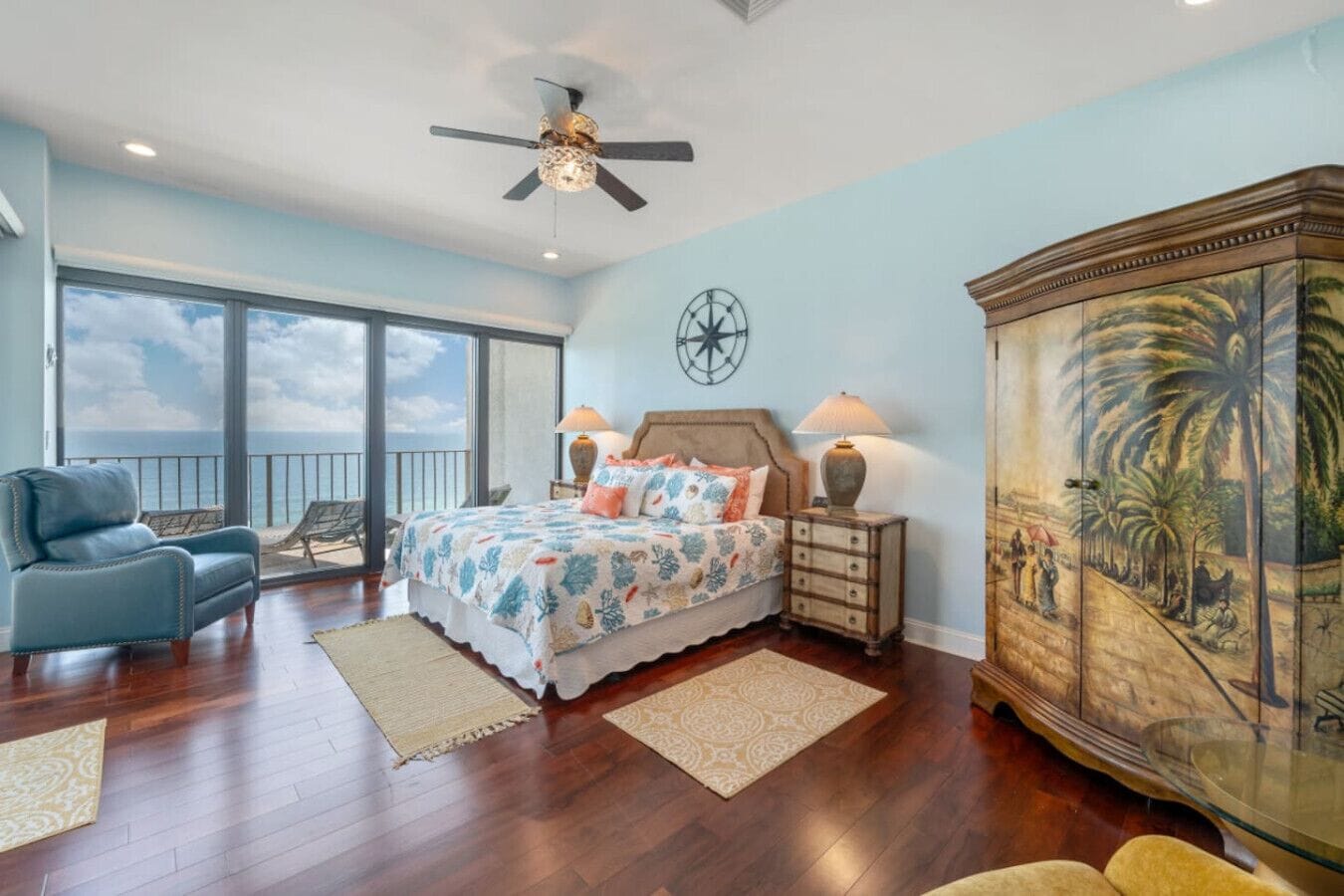 Une chambre spacieuse avec un grand lit, un ventilateur de plafond, une table de chevet avec des lampes, un fauteuil bleu et une vue sur l’océan grâce à de grandes fenêtres.