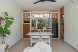A modern kitchen with white cabinetry, a dining table, and chairs. There is a large window with a view of a residential area. - Dreamscape Haven  (Canggu)