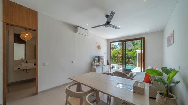 A modern dining area with a wooden table and chairs, a ceiling fan, and a view of a pool and greenery outside. - Dreamscape Haven  (Canggu)