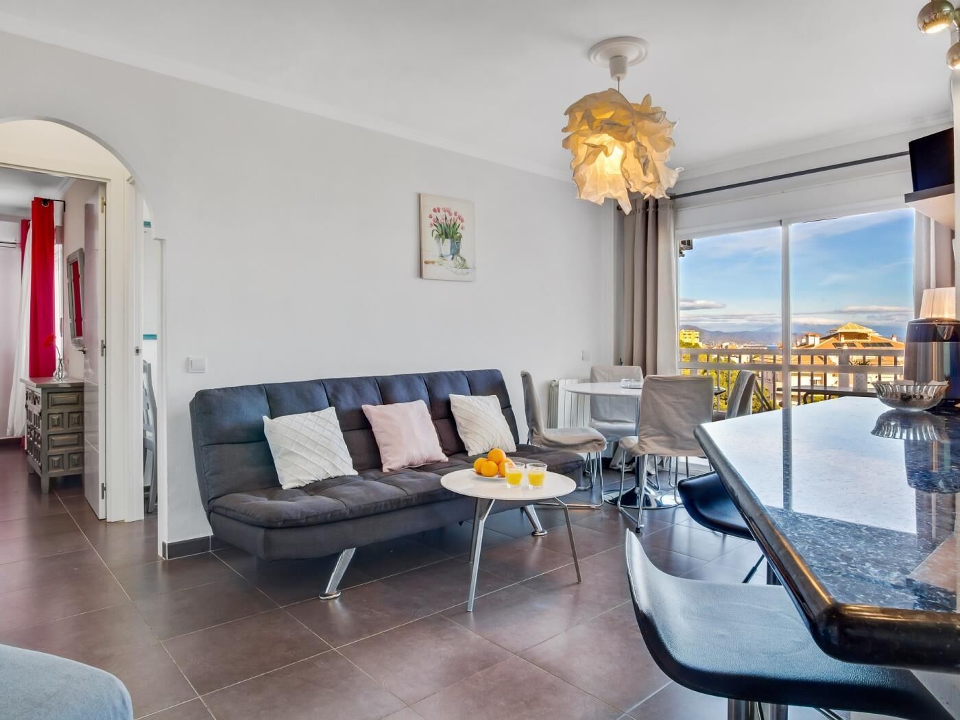En moderne stue med en mørkegrå sofa, et rundt sofabord med appelsiner og juice, et spiseområde med bar, og et stort vindue med udsigt over byen.