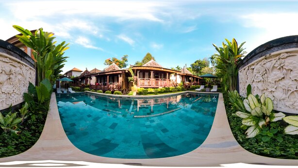 A swimming pool with a curved edge, surrounded by tropical plants and a building with a thatched roof. - Ari Cottages (Ubud)
