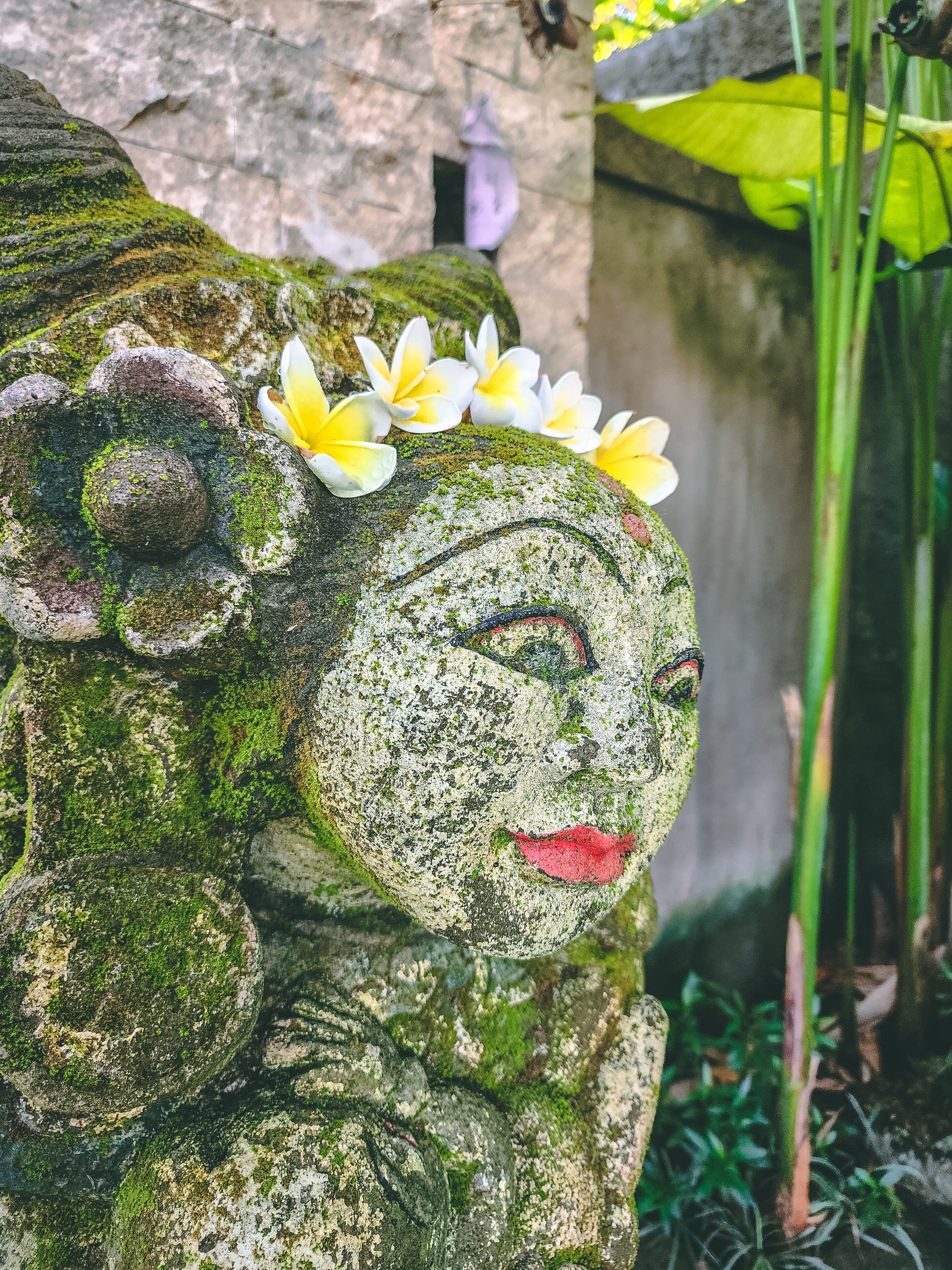 A stone statue with a floral headpiece, moss-covered, and set against a stone wall with greenery in the background.