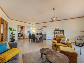 A living room with a sofa, armchair, coffee table, dining table, and chairs. There is a potted plant, a painting, and a bookshelf. - Aguia M T2 - VIP Properties by Interhome (Albufeira)