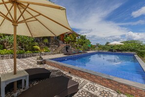 A rectangular swimming pool with blue tiles, surrounded by a brick border and a tiled patio. There are lounge chairs and a table under a large umbrella. - La Village Hill View  (Penida Island)