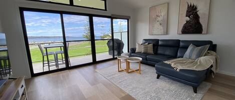A spacious living room with a large sliding glass door leading to a balcony, a dark blue sofa, a small round coffee table, and two wall-mounted paintings.