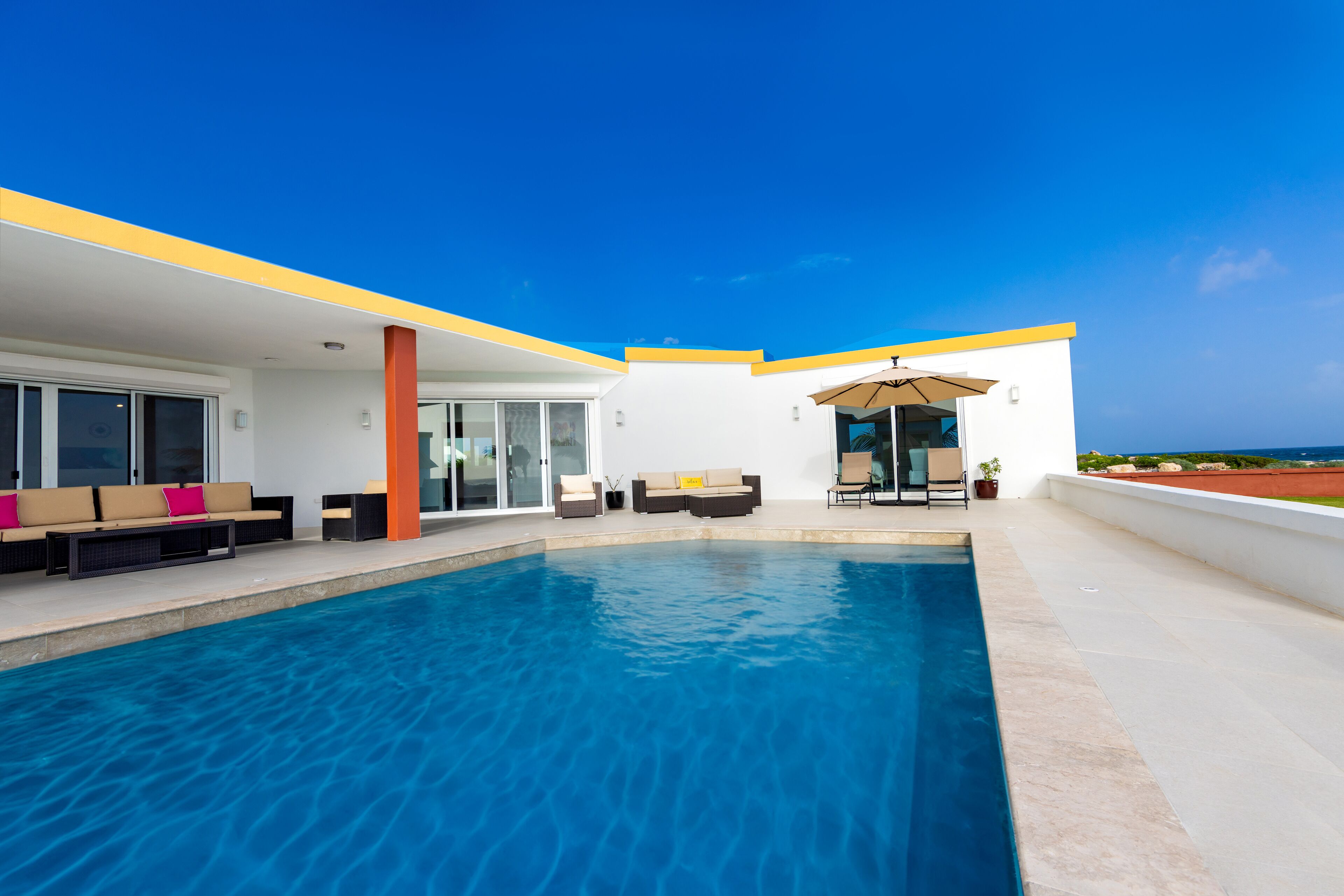 A modern outdoor swimming pool area with a clear view of the ocean, a covered seating area, and a lounge with sofas and chairs.