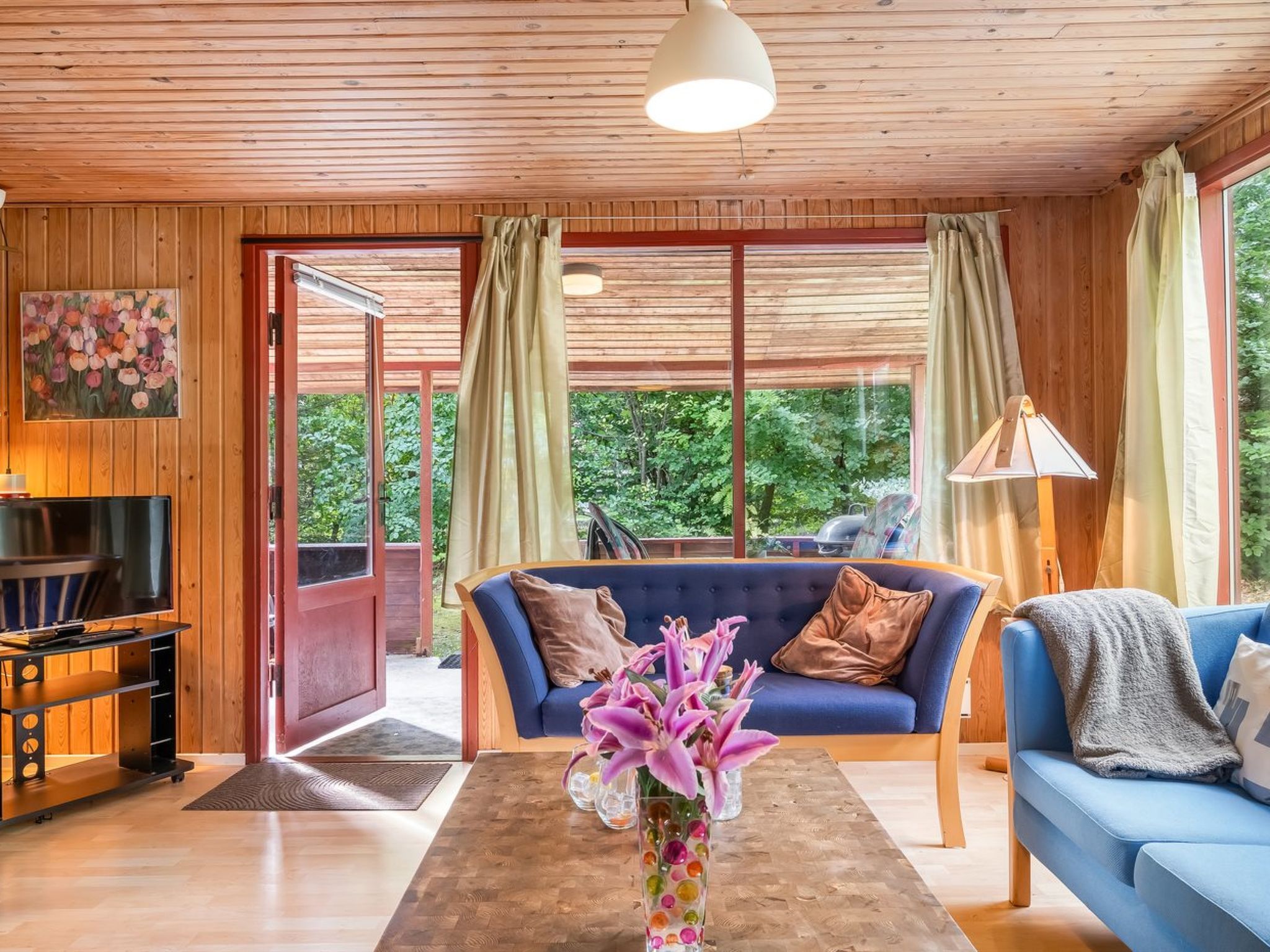 A cozy living room with a blue sofa, a coffee table with a vase of flowers, a television, and a large window with curtains.