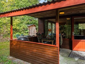 A wooden cabin with a covered patio area, a grill, and a dining table with chairs.