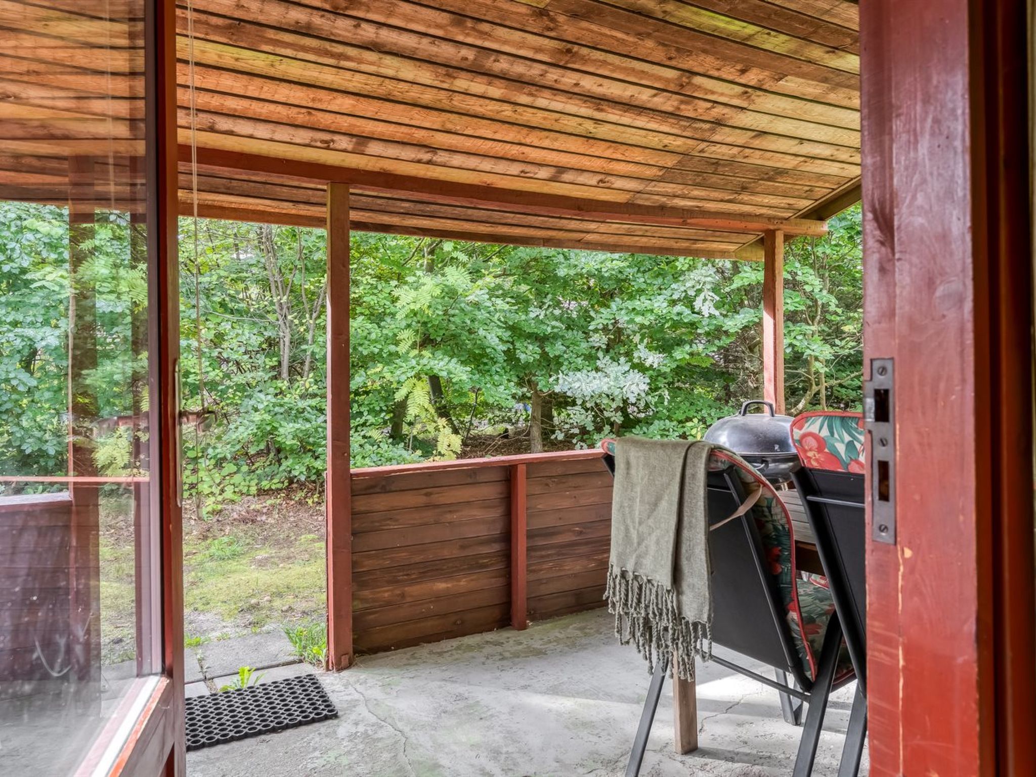 Un espace extérieur abrité, équipé d’un barbecue, d’un banc et offrant une vue sur une zone boisée.