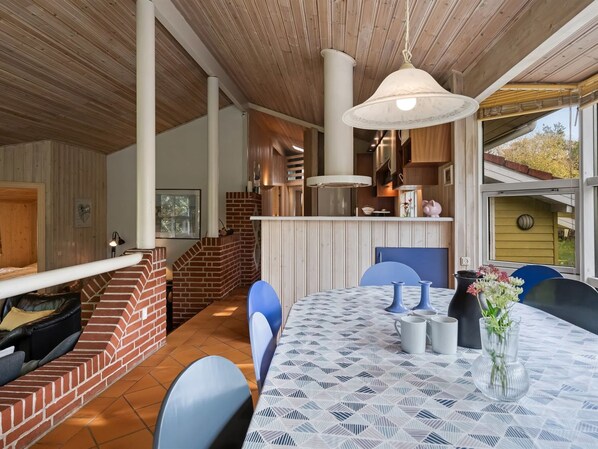 A dining area with a table set for two, a brick fireplace, and a kitchen in the background.