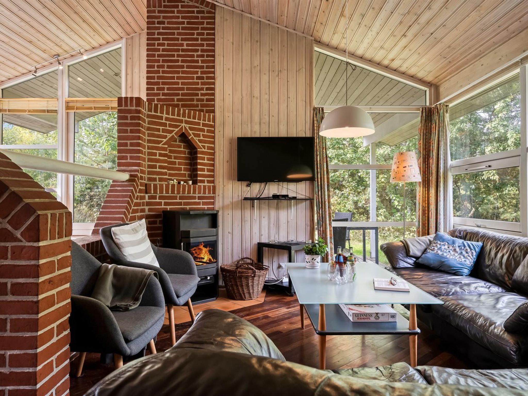 A cozy living room with a fireplace, comfortable seating, a flat-screen TV, and large windows offering a view of the outdoors.