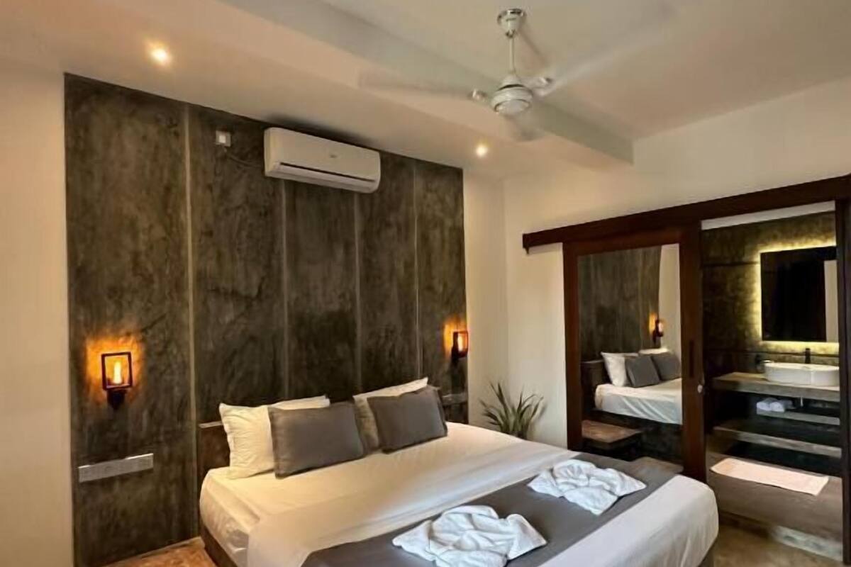 Quarto moderno com cama grande, ar condicionado embutido, ventoinha no teto e banheiro com espelho amplo.