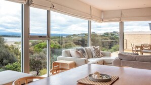 A spacious living area with a large window overlooking a beach and greenery, a wooden dining table, and a sofa with cushions. - Sea View - Freycinet Holiday Houses (Coles Bay)