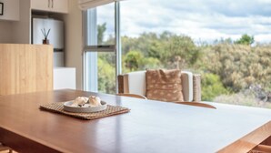 A modern kitchen with a wooden table, a bowl of food, and a view of a forest through large windows. - Sea View - Freycinet Holiday Houses (Coles Bay)