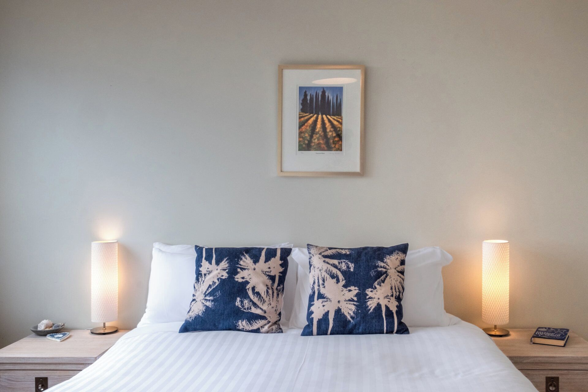 Una cama bien hecha con ropa blanca y dos almohadones decorativos, flanqueada por mesitas de noche con lámparas y un cuadro enmarcado encima.