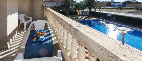 A balcony with a pool, white plastic chairs, a table with a blue tablecloth, and a view of the city.