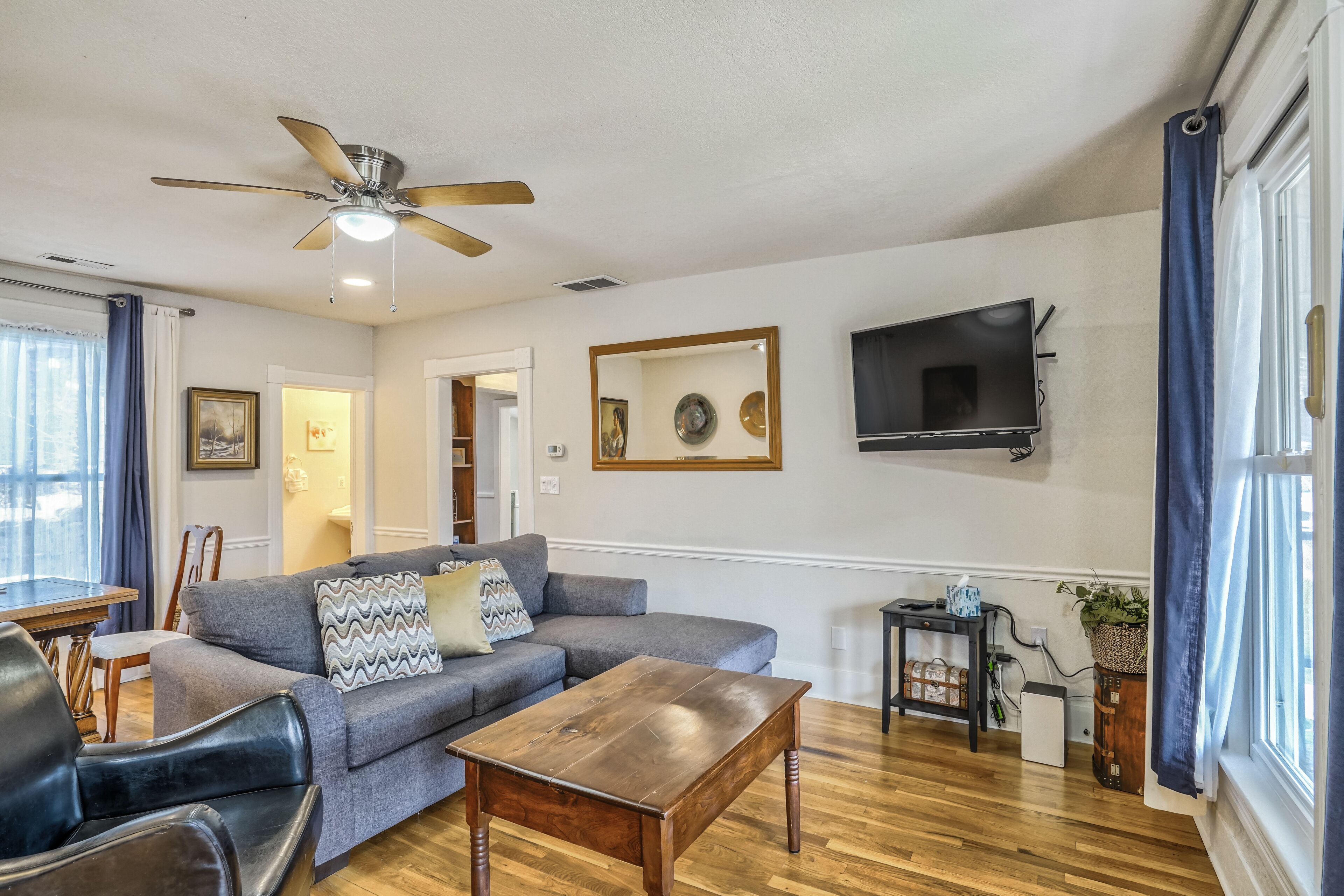 A living room with a grey sofa, wooden coffee table, ceiling fan, flat-screen TV, and a small table with a plant.
