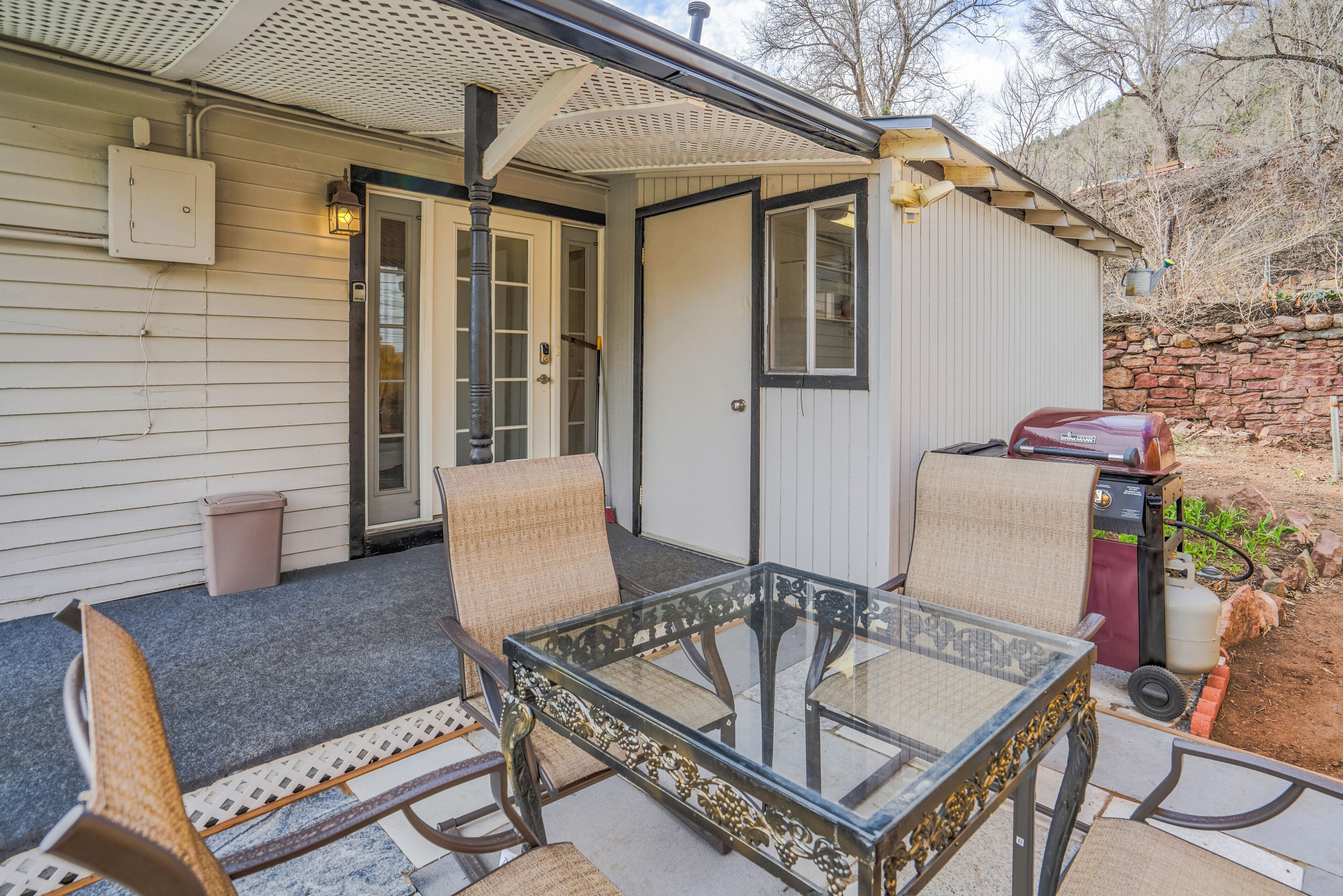 Une terrasse couverte avec une table en verre et quatre chaises, un barbecue et un mur de pierres en arrière-plan.